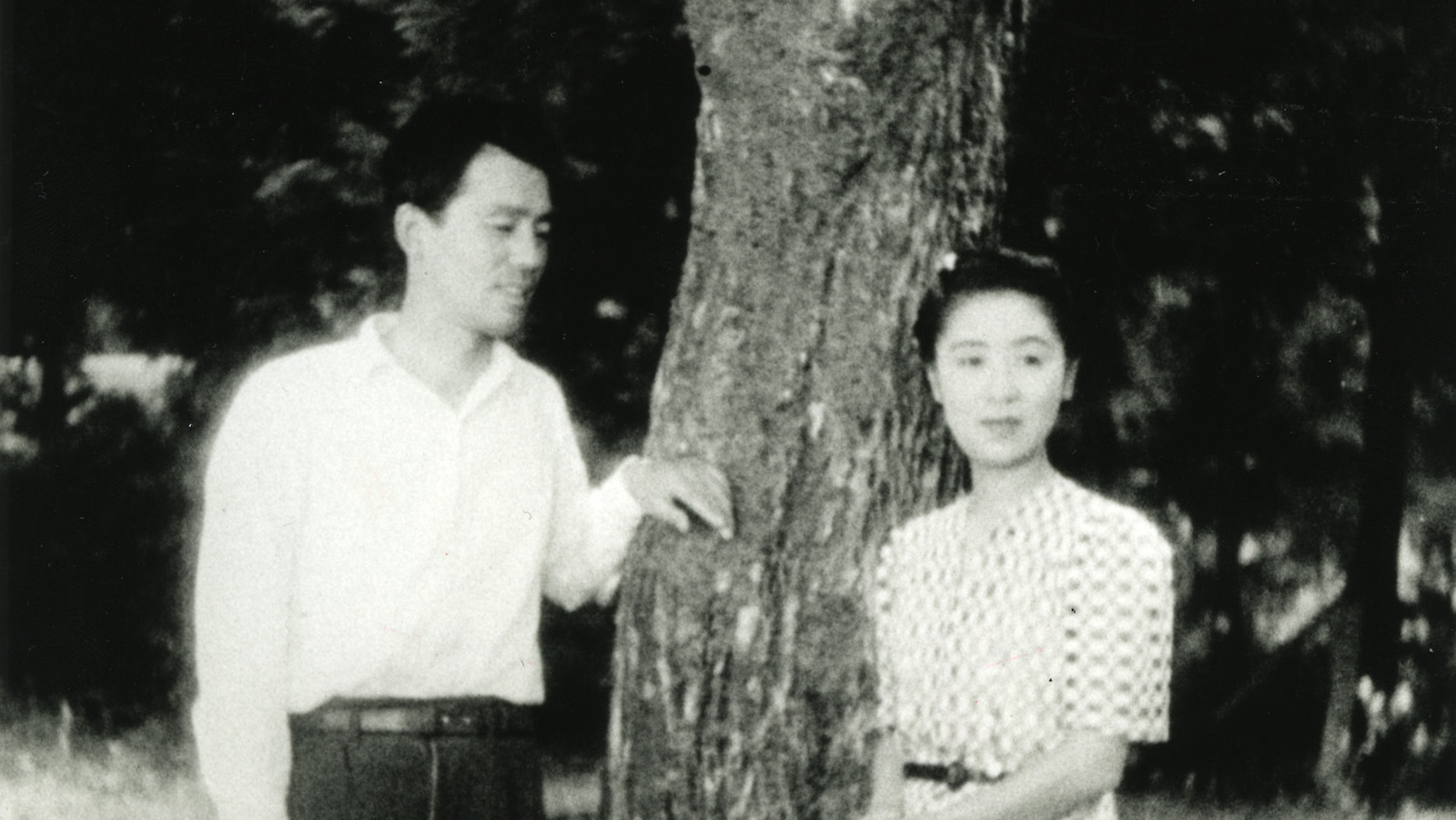 A black and white photo of a Japanese man and woman standing by a tree outside; he looks at her, she looks off past the camera.
