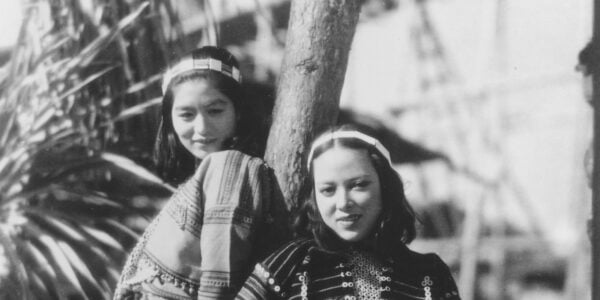A black and white photo of two Japanese women leaning against a wooden pole and looking hopefully into the camera from a high angle.