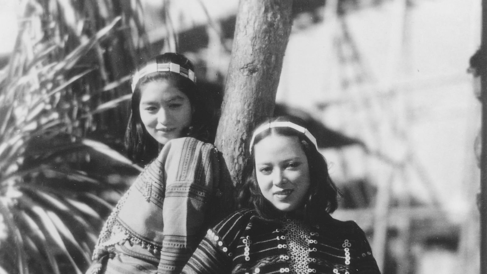 A black and white photo of two Japanese women leaning against a wooden pole and looking hopefully into the camera from a high angle.