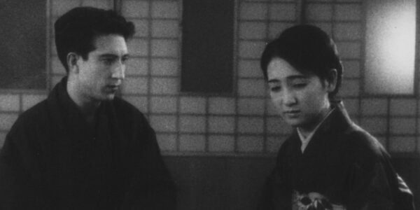 black and white image of a young Japanese couple, the man looking down at a woman who looks away, both sitting in front of a traditional screen