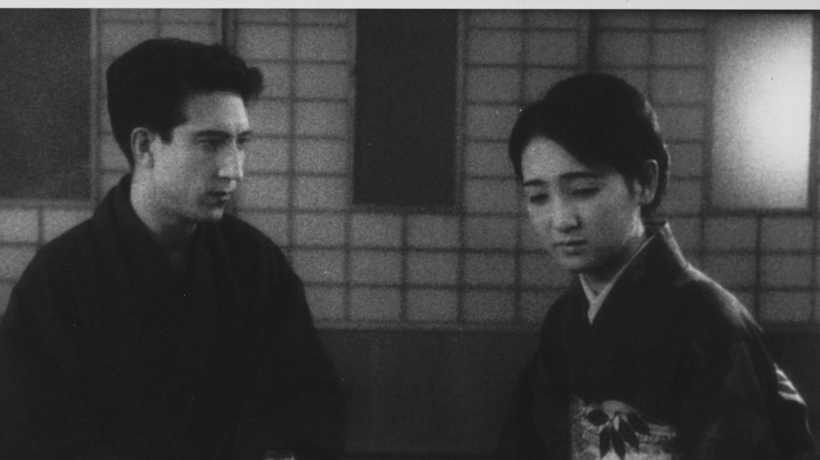 black and white image of a young Japanese couple, the man looking down at a woman who looks away, both sitting in front of a traditional screen