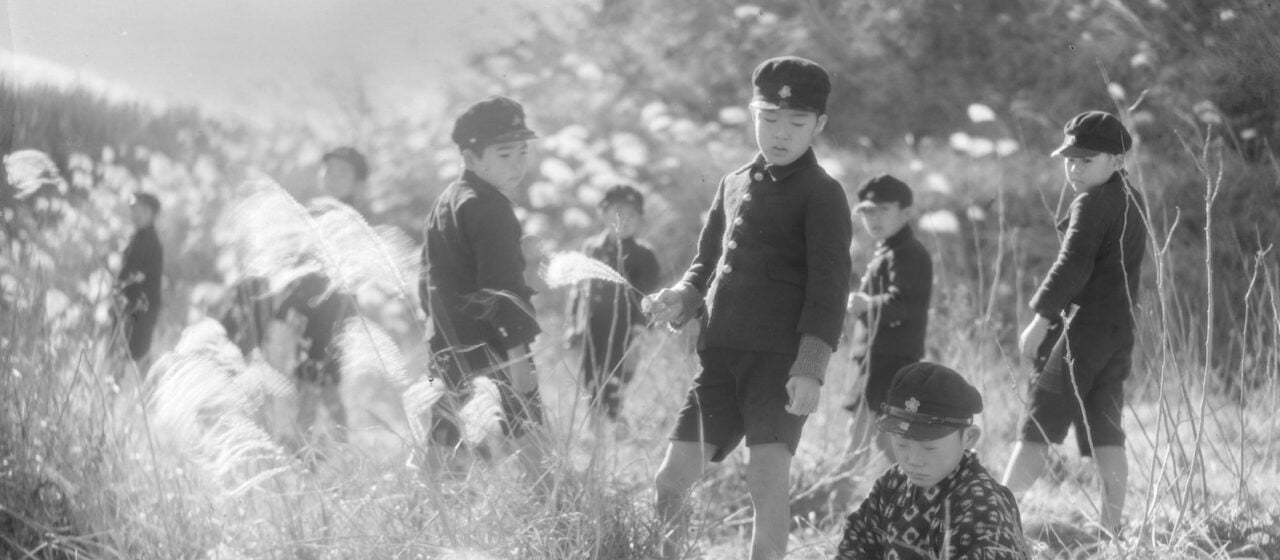 A black-and-white image from the 1930s of young Japanese boys dressed in school uniforms and caps standing amidst a sun-dappled field of wheat