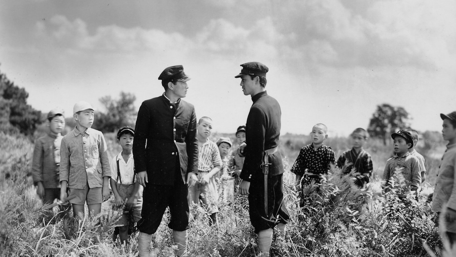 A black and white photo of two men in hats and uniforms standing and facing each other in a field of grass amidst other children figures who surround and watch them.
