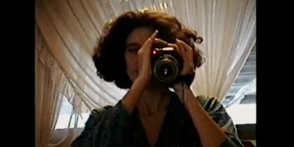 An image that looks like slightly degraded video from the eighties or nineties in which a woman holds a camcorder to her eye and aims toward the screen.