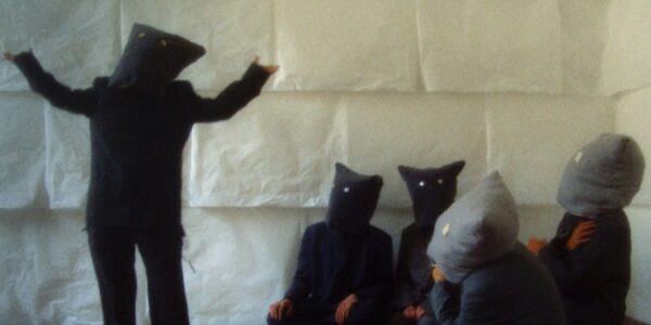 A group of five people with bags over their heads and buttons for eyes in a room with white walls. One of the people stands with their arms outstretched and the others are sitting on the floor