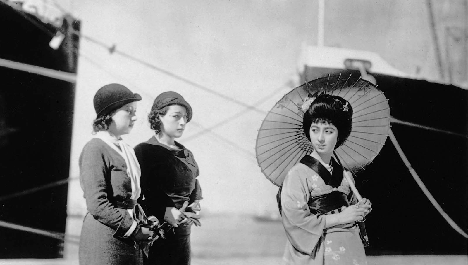At a seaside dock, two Japanese women in modern clothes and hats look at a woman in traditional geisha clothes and parasol