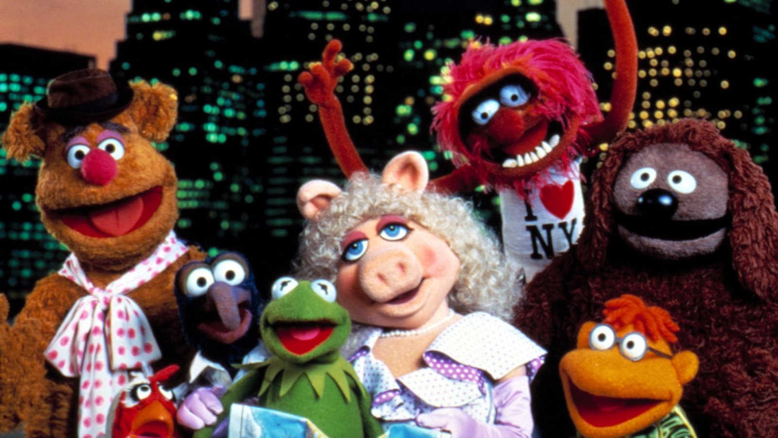 A number of colorful puppets, best known as Muppets, stand in front of a night-time New York City skyline.