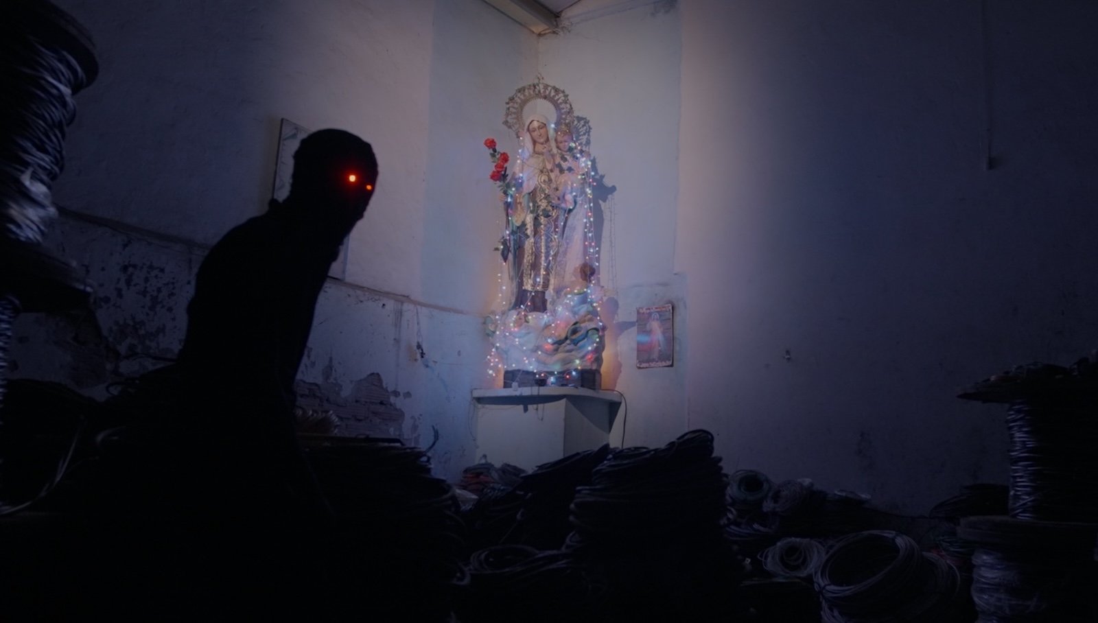 A dark figure with glowing red eyes stalks a room at night, where a Virgin Mary statue sits in the corner holding a rose.