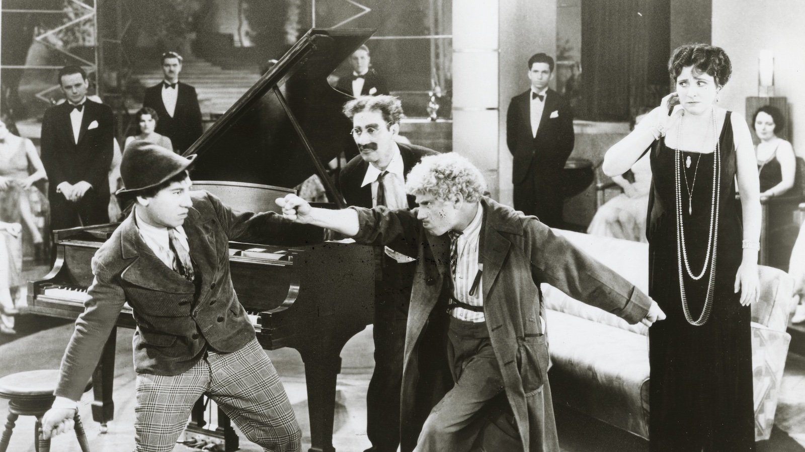 Two men mock fight in front of a grand piano at a party while a man with a painted-on mustache eggs them on and a woman in an evening gown and long pearl necklace watches from the sides.