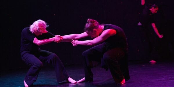 Bathed in purple light, two dancers grasp each other's hands and pull on their arms, with their heads down, amidst a black-box space