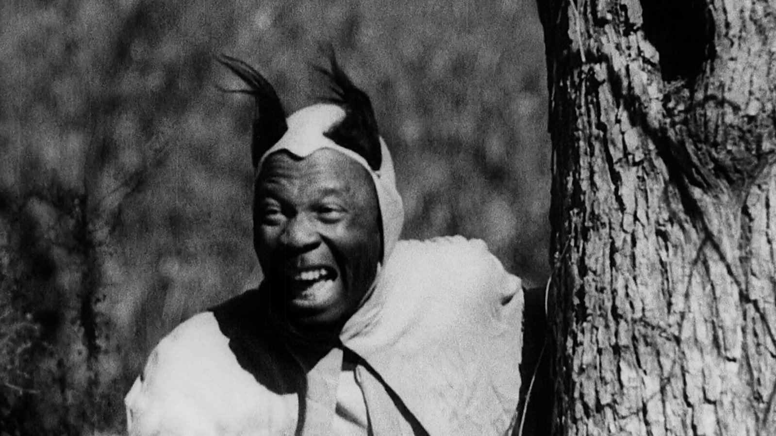 A medium brown skinned man in a devil costume with horns peers from behind a tree with his mouth open in a diabolical laugh