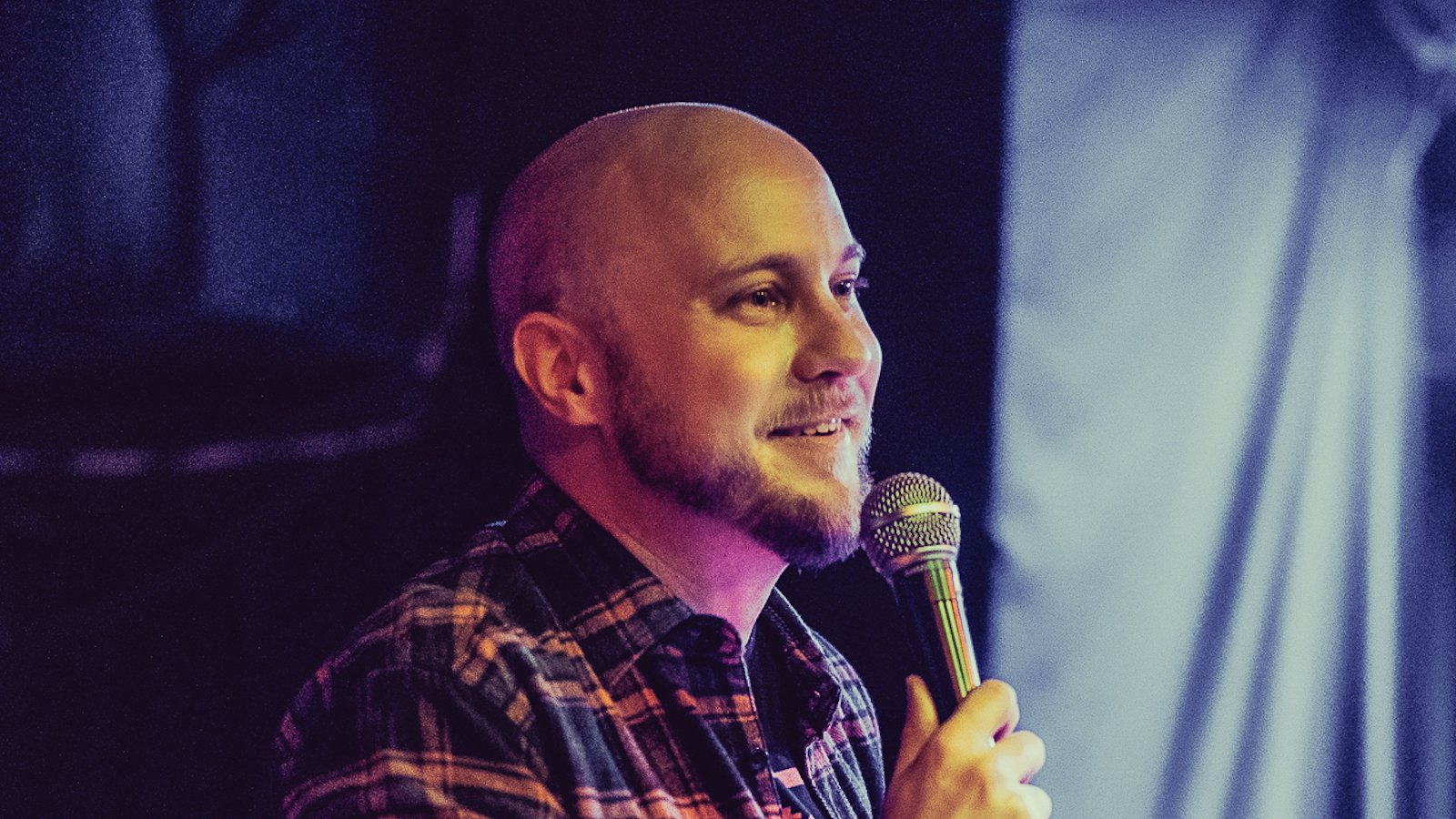 A bald man with a beard smiles and wearing a plaid shirt talks into a microphone
