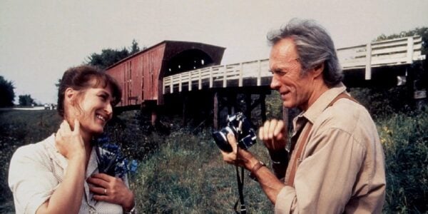 Standing in front of a covered bridge in a rural setting, a man holds a camera, pointing a lens towards a woman who blushes as she grabs her neck.