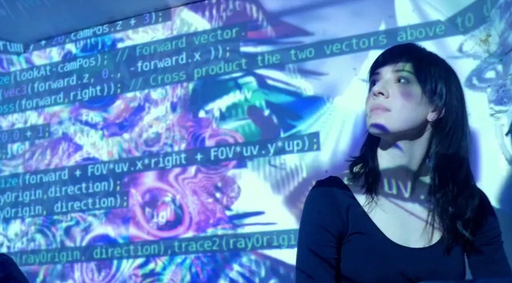 A woman in long dark hair stands against a wall against which computer codes and abstract purple and blue images are projected