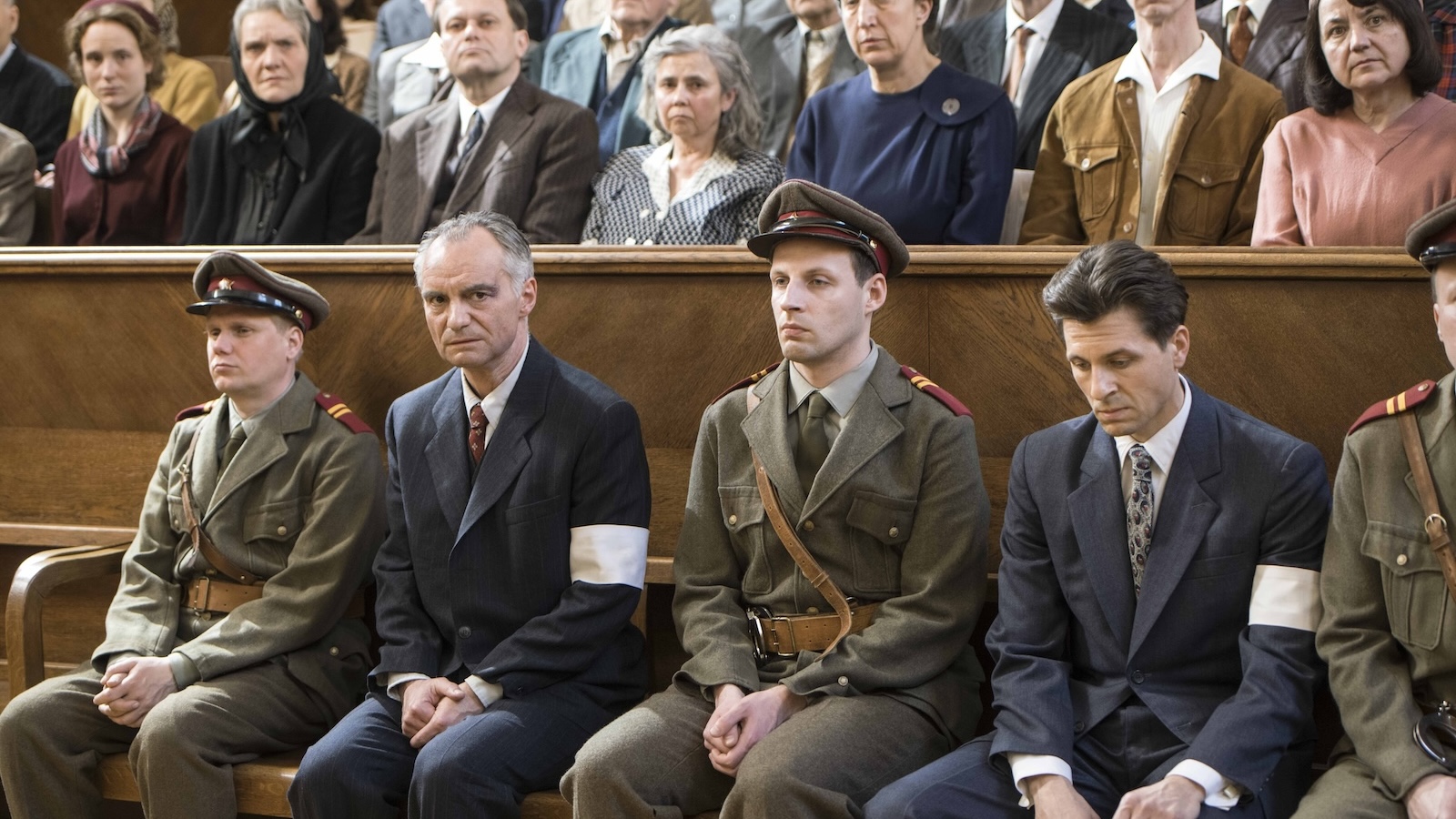 A row of men in a courtroom, who appear to be the defendants on trial, two of them wear blur suits and white armbands, the other two are dressed in military uniforms, all in WWII-era outfits