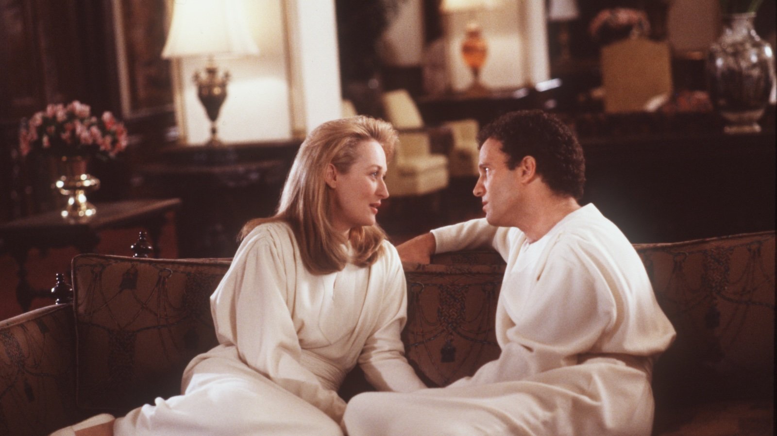 A man and woman in matching white robes sit on a couch