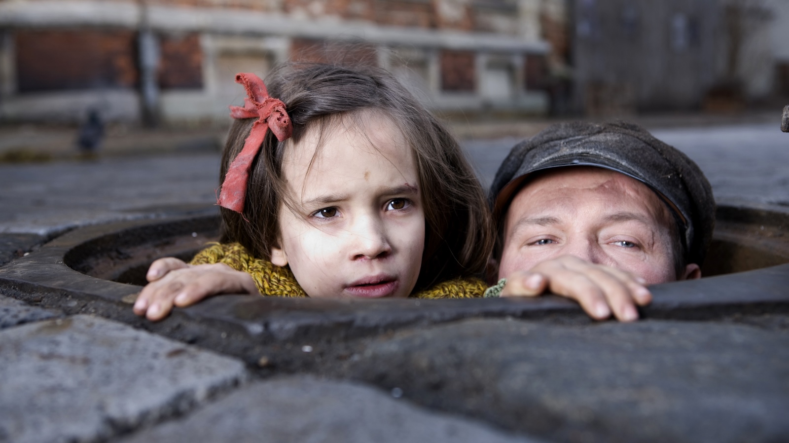 Two children, the girl with a red ribbon in her hair and a boy with a cap, poke their heads out of a sewer grate