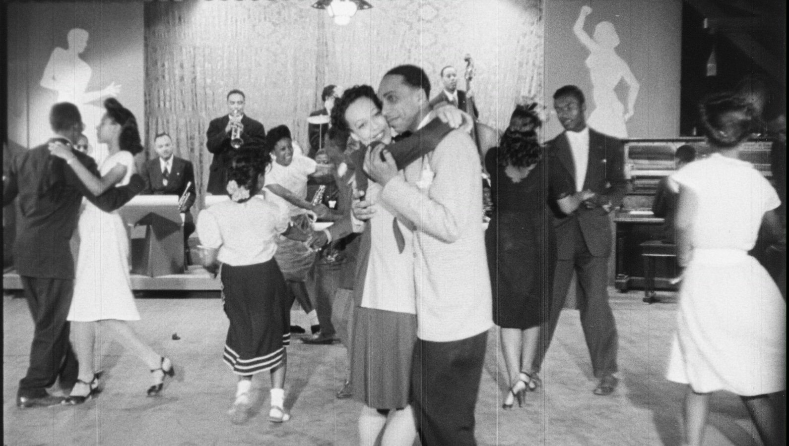 An African American woman and man dance together cheek to cheek amidst other dancers at a juke joint in this black and white image from the 1940s.