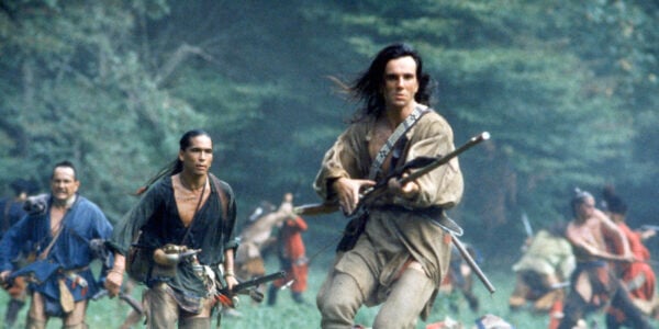A long-haired man with a rifle, surrounded by other Native American warriors, runs through a field surrounded by trees and mountains