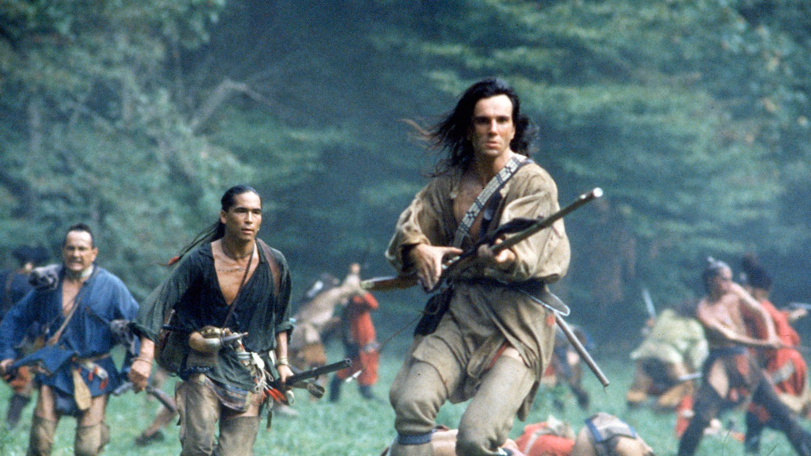 A long-haired man with a rifle, surrounded by other Native American warriors, runs through a field surrounded by trees and mountains