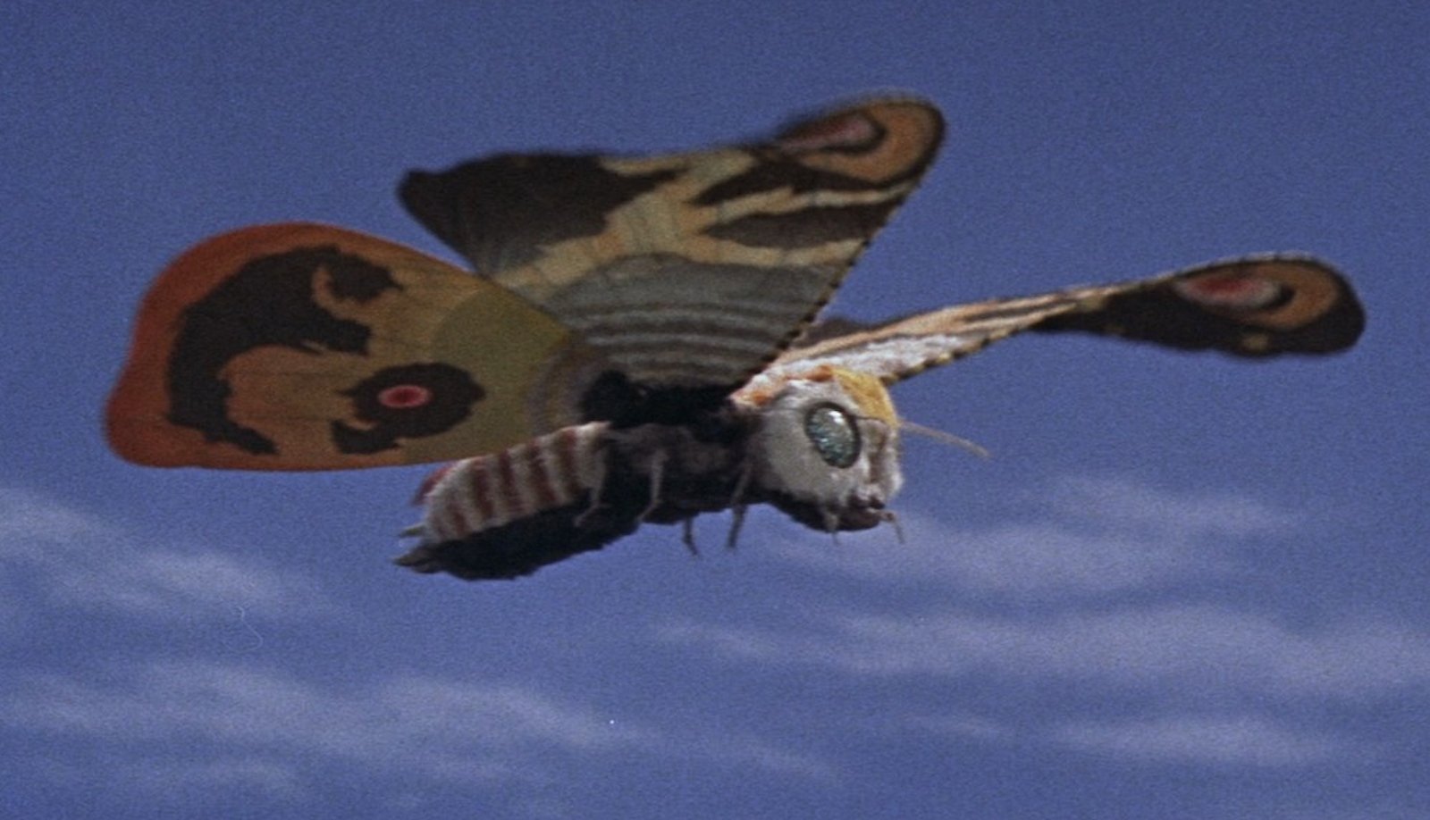 A multicolored moth, clearly fake, flies in the air against a blue sky