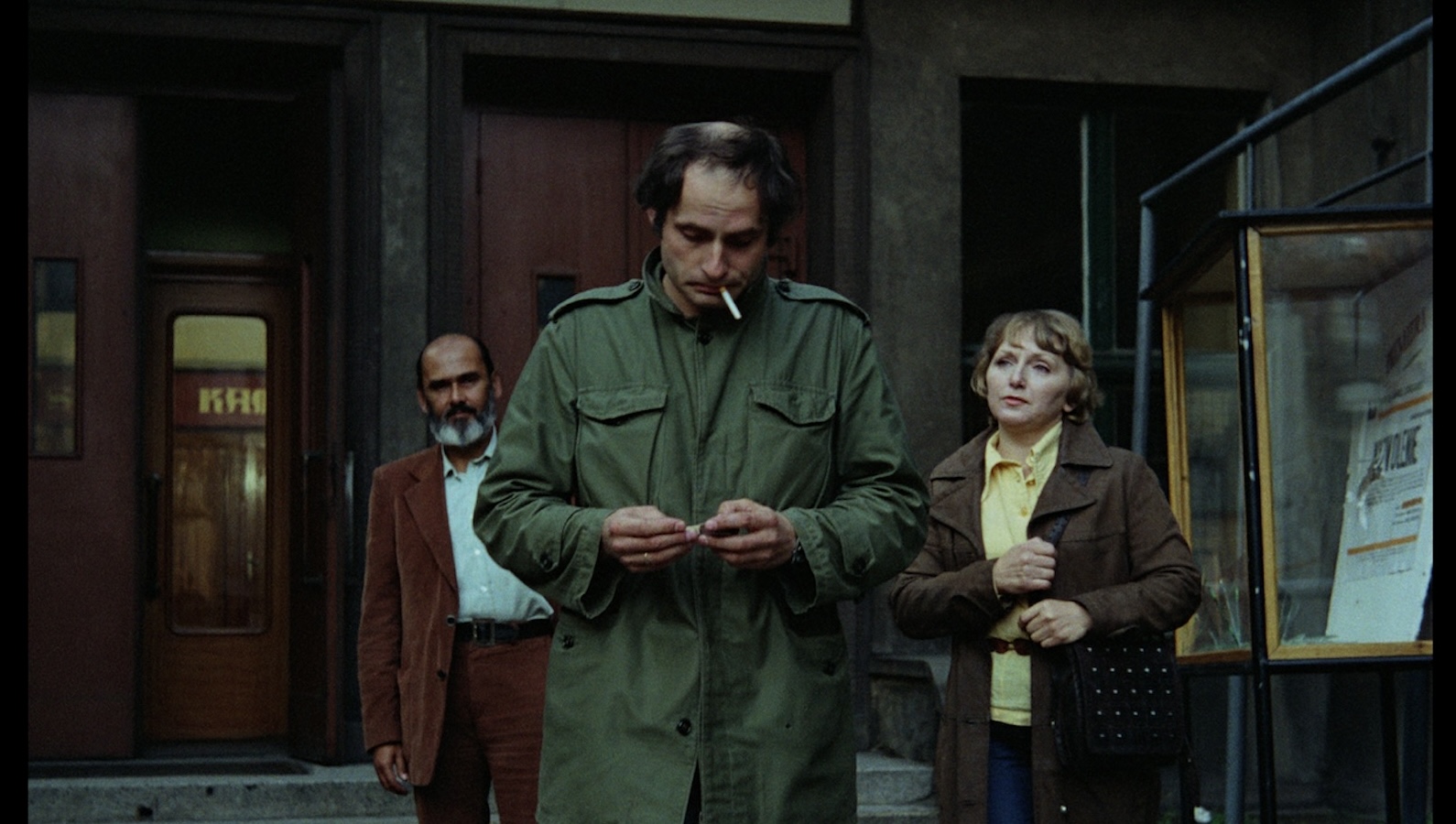 A man in green fatigues lights is about to light a cigarette dangling from his mouth, while a man and woman walk behind him, looking expectantly at him