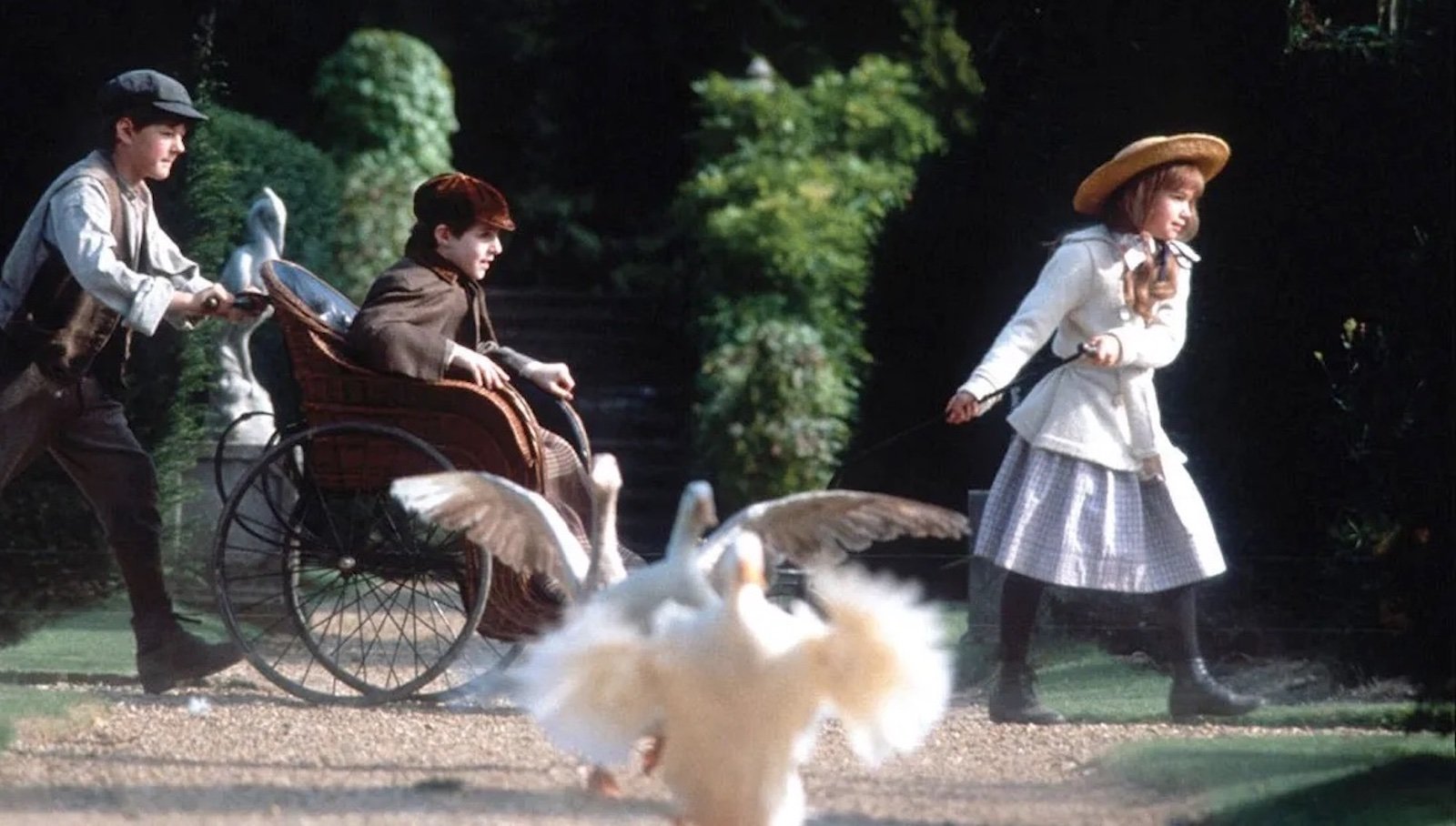 In an old English garden setting, a boy in a cap pushes a wheelchair with a boy in an cap sitting in it, as the whole chair is pulled along in front by a girl in a straw hat. In the foreground are geese flapping their wings.