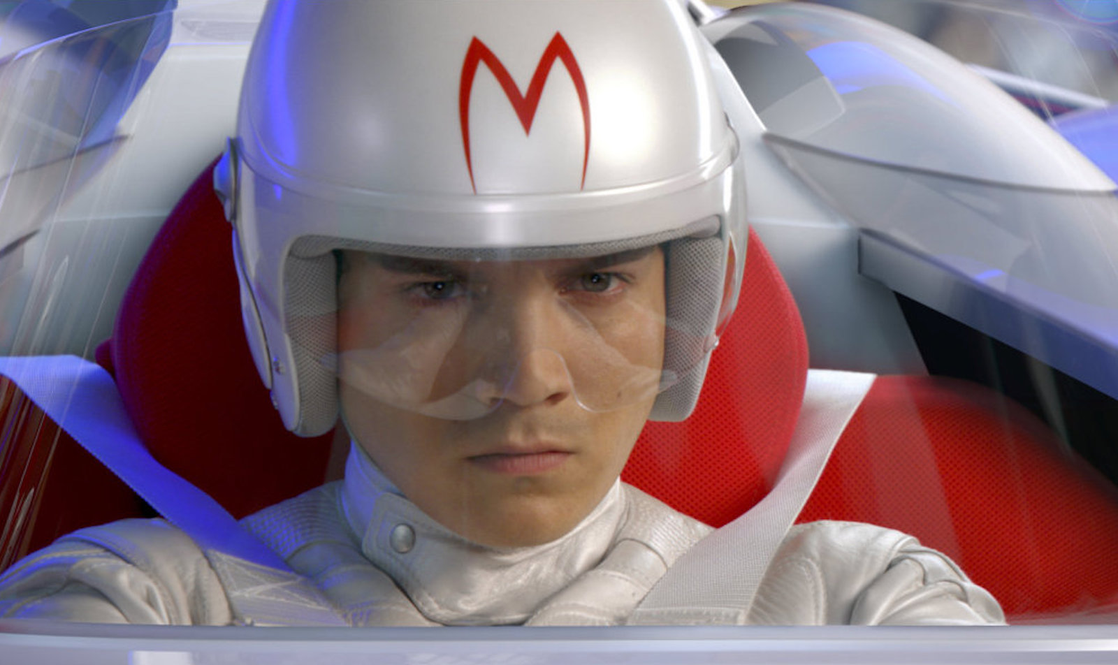 A serious-looking car racer in goggles and white helmet with a red M painted on the front is strapped into his vehicle, looking straight ahead with determination.