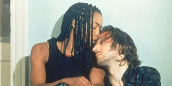A brown-skinned woman in dreadlocks kisses the forehead of a light-skinned man with a bloody forehead as they crouch against a white wall