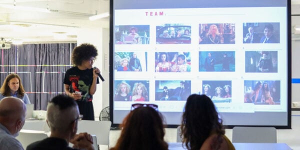 A woman with curly hair speaks into a mic next to large screen showing small boxes of images from films as part of a trivia game; in the front, the backs of participants heads can be seen looking at the screen.