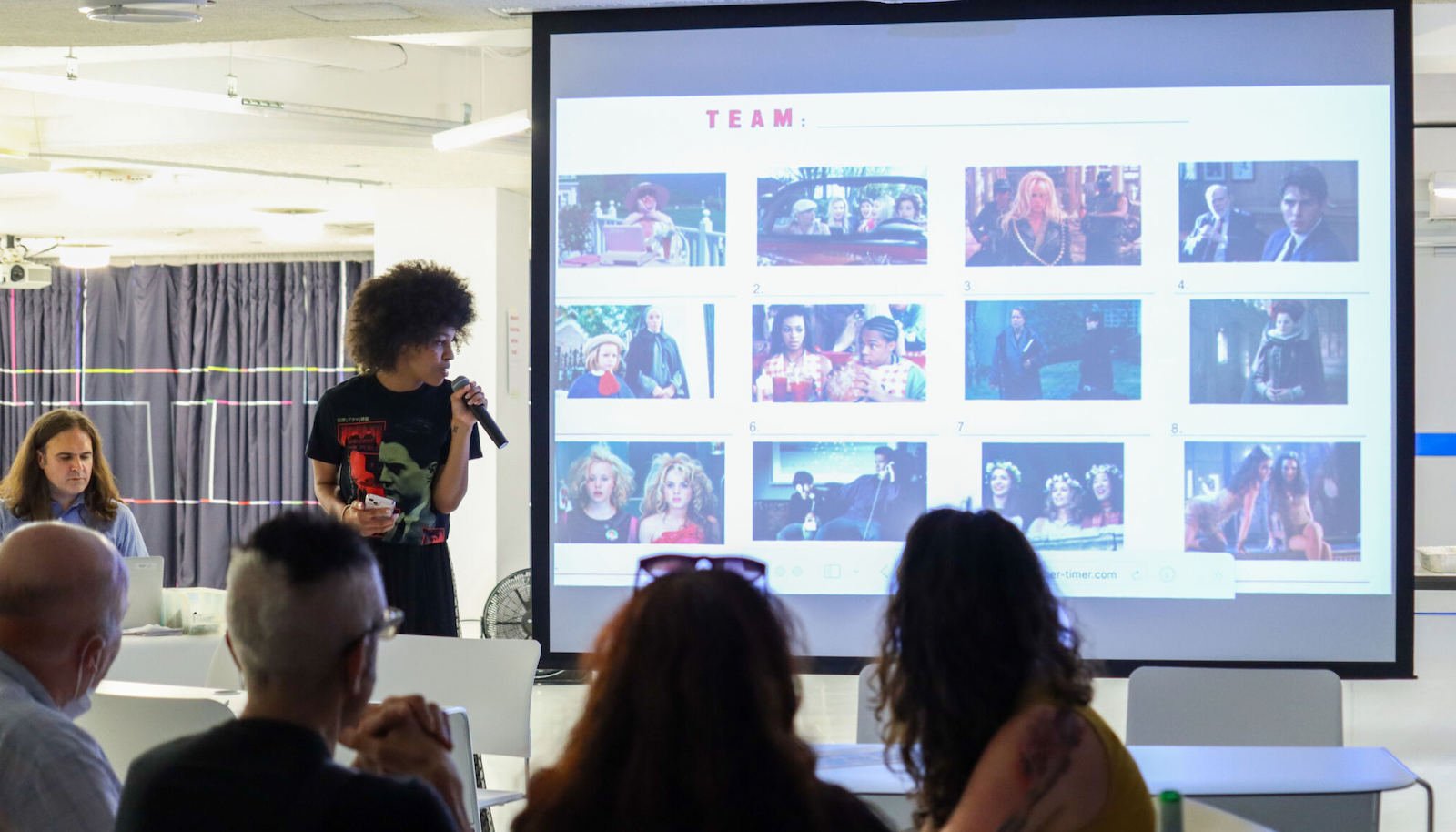 A woman with curly hair speaks into a mic next to large screen showing small boxes of images from films as part of a trivia game; in the front, the backs of participants heads can be seen looking at the screen.