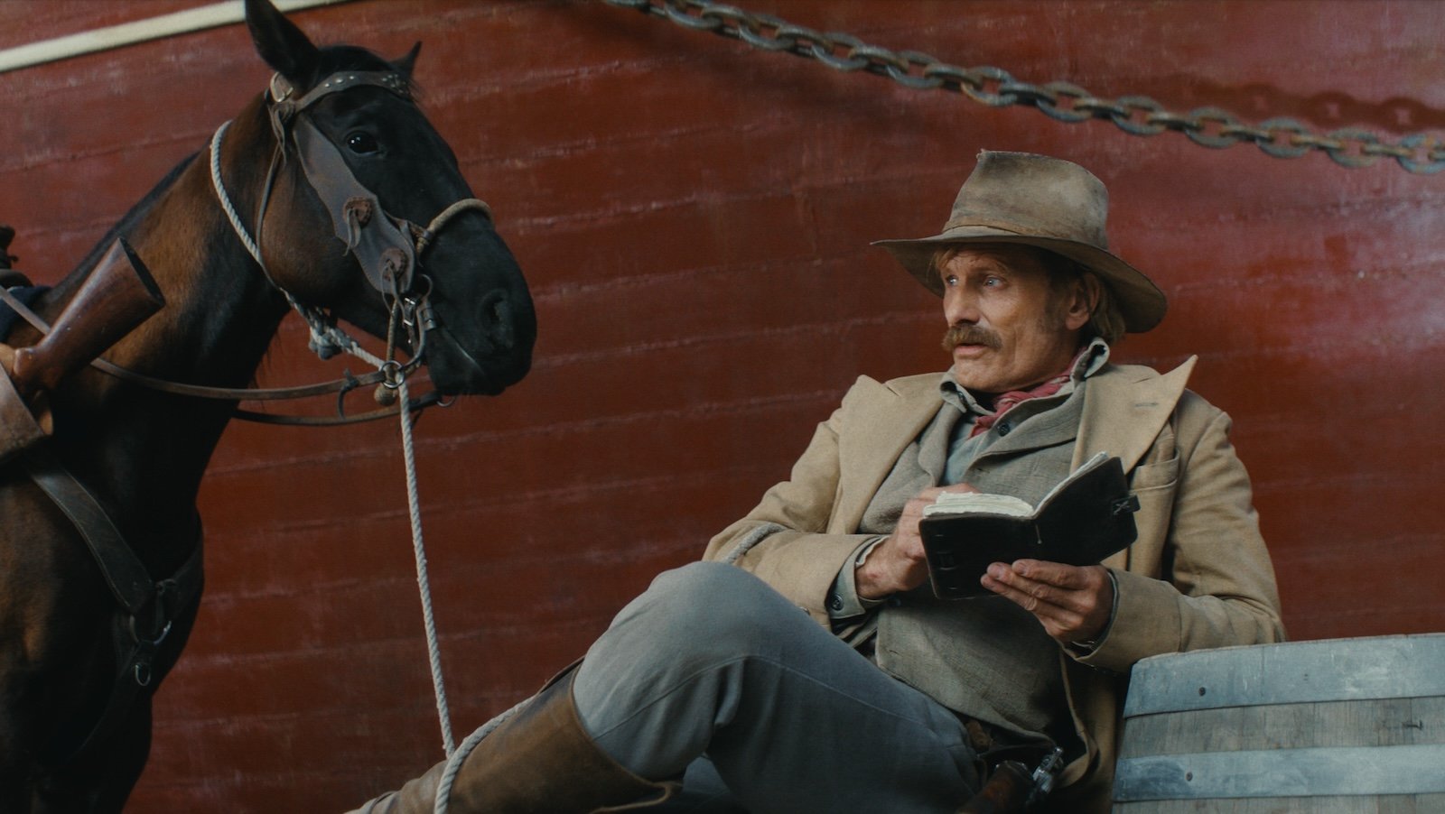 A man in a fedora sits with his legs crossed reading a book leaning against a red brick wall; a horse stands beside him.