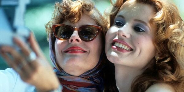 Two women smile take a polaroid of themselves, both wearing makeup, the one on the left in sunglasses and kerchief.