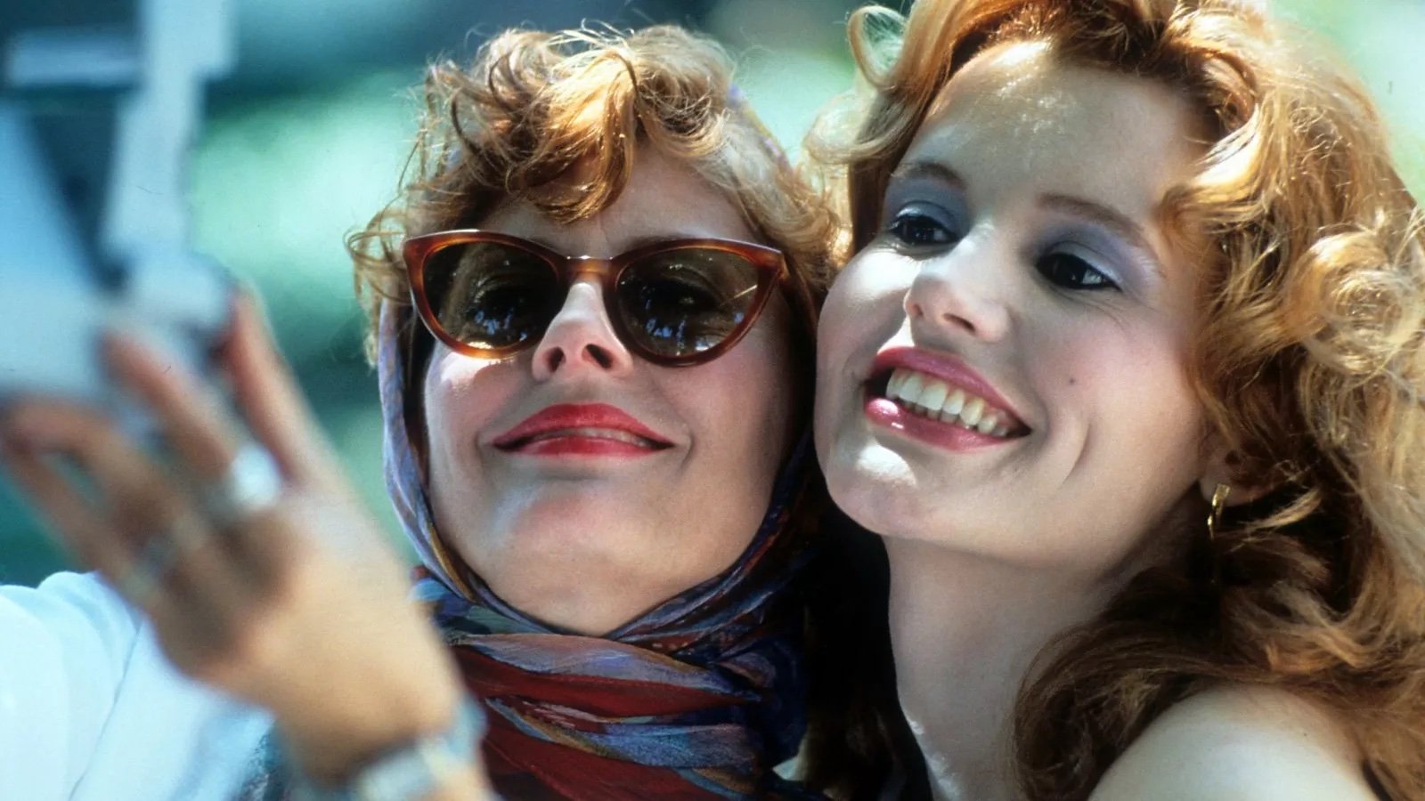 Two women smile take a polaroid of themselves, both wearing makeup, the one on the left in sunglasses and kerchief.