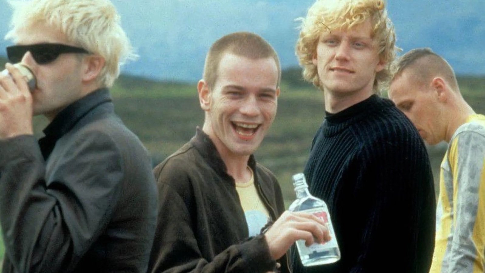 A group of four pale-skinned young men stand in a field, the second from the left is smiling as he holds an open bottle of liquor