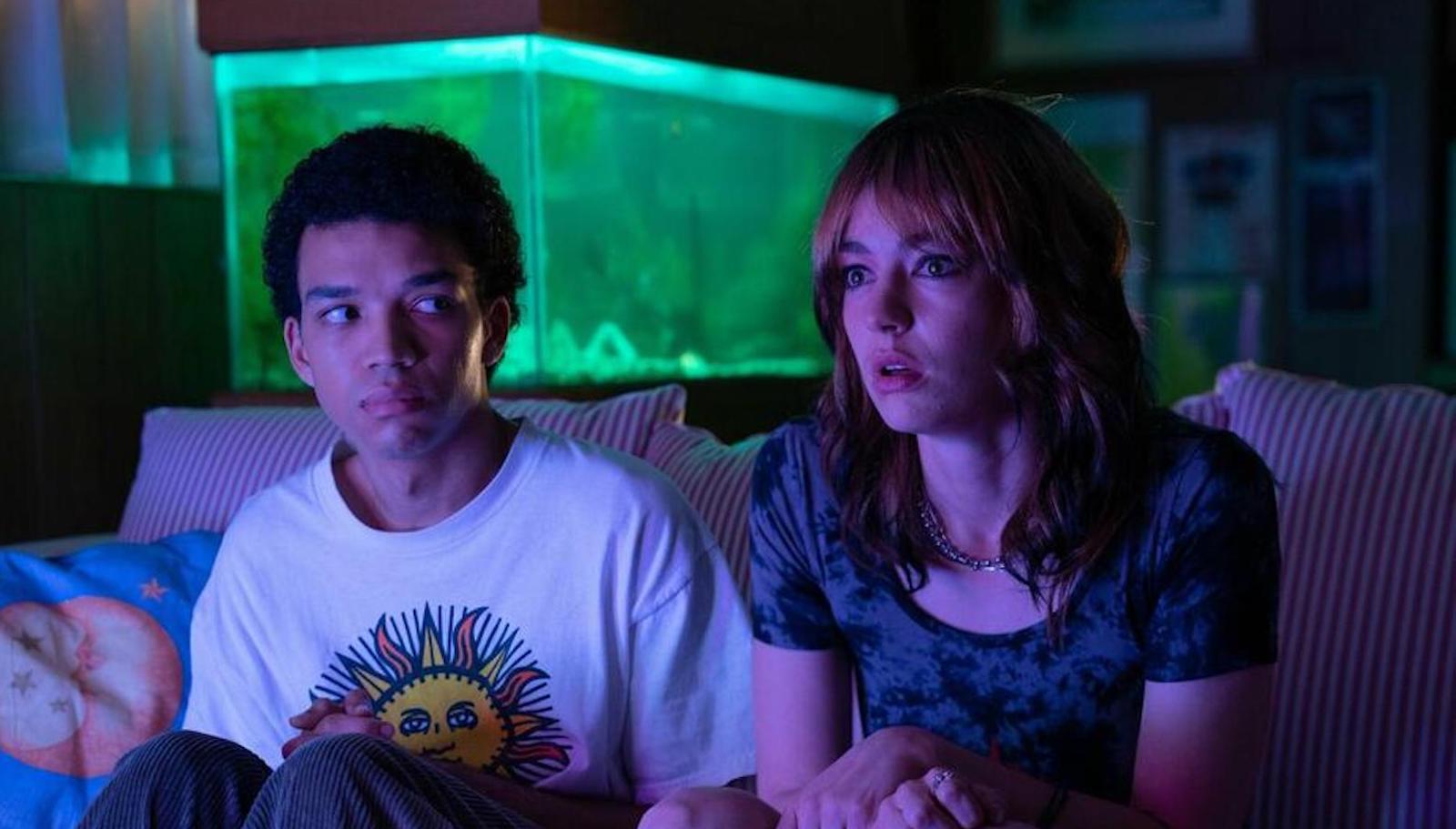 Two teenagers, one with short hair and medium brown skin and one with long hair and pale skin and red long hair, sit on a couch with a purple TV glow cast on them and a green light background.