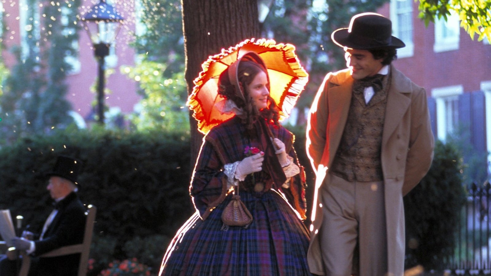 A woman with a yellow and orange parasol lit up by the sun walks down a city street next to a man in a hat; they are smiling and are wearing late nineteenth-century-style clothes