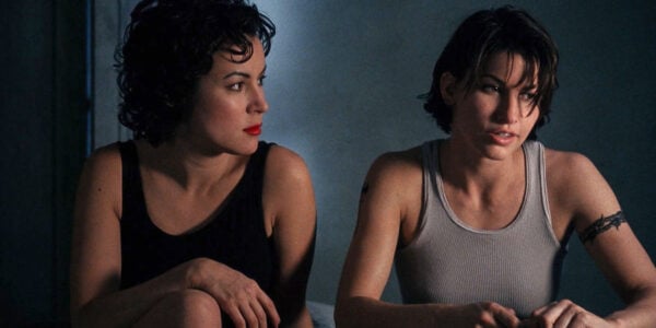 Two women, one traditionally feminine in a black dress and lipstick and the other more butch in a gray tank top sit next to each other in a dark room