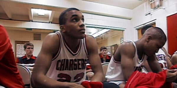 A young man of medium brown skin wearing an athletic tank top with the word CHARGERS on it sits in a locker room and looks pensively.