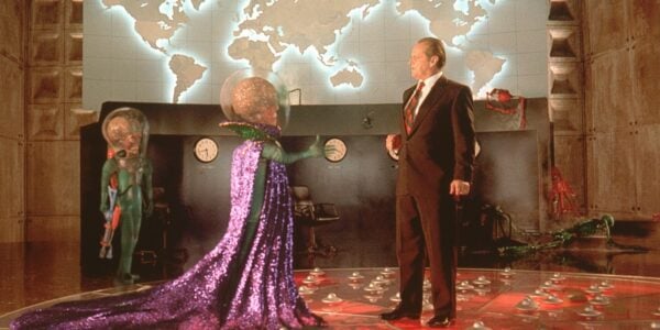 A silly-looking skeleton-faced martian with a huge brain reaches out its hand to shake the hand of a hesitant human man in a brown suit with a map of the world behind them