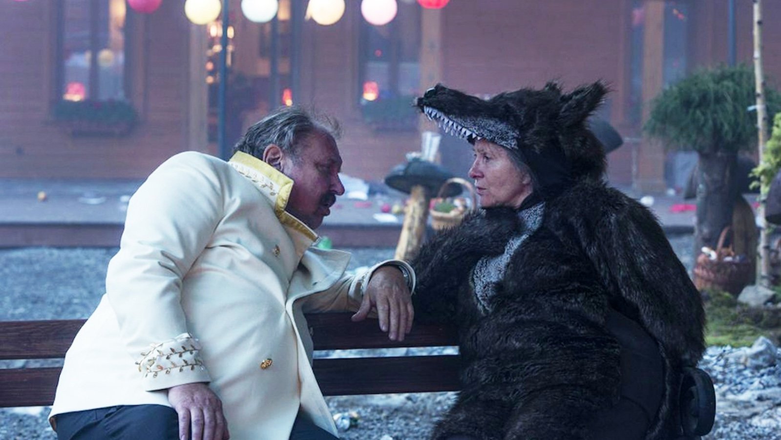 Two people sit on a bench looking at one another: the man is in a white jacket, the woman is wearing a wolf costume