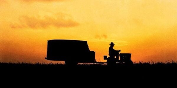 A silhouetted tractor rides down a road at twilight against on orange sky, with a human figure at the front.