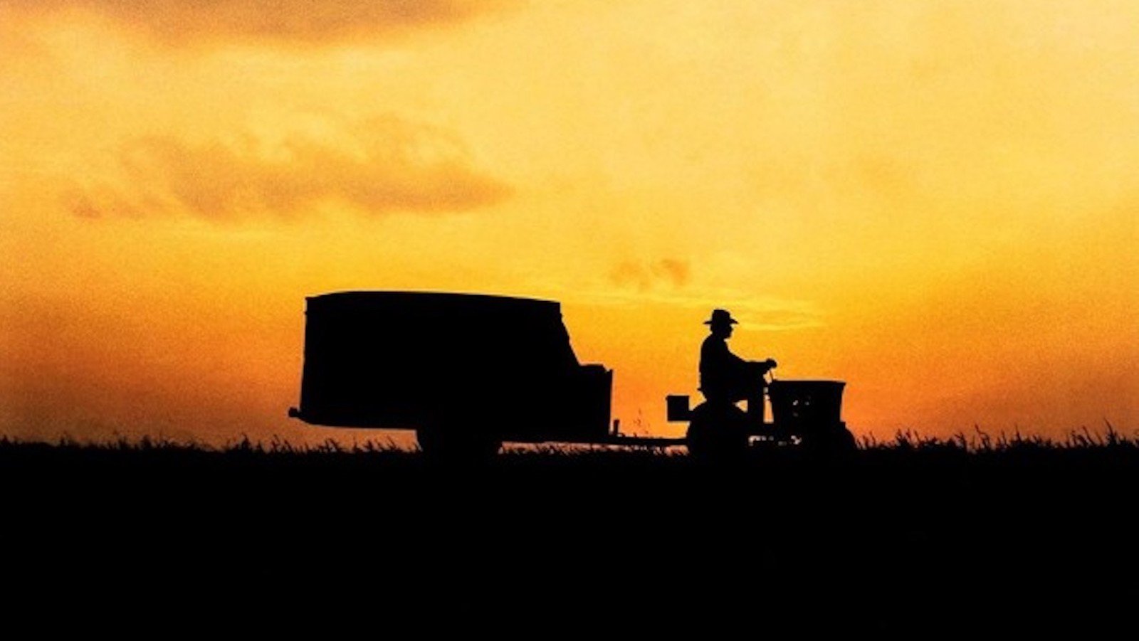 A silhouetted tractor rides down a road at twilight against on orange sky, with a human figure at the front.