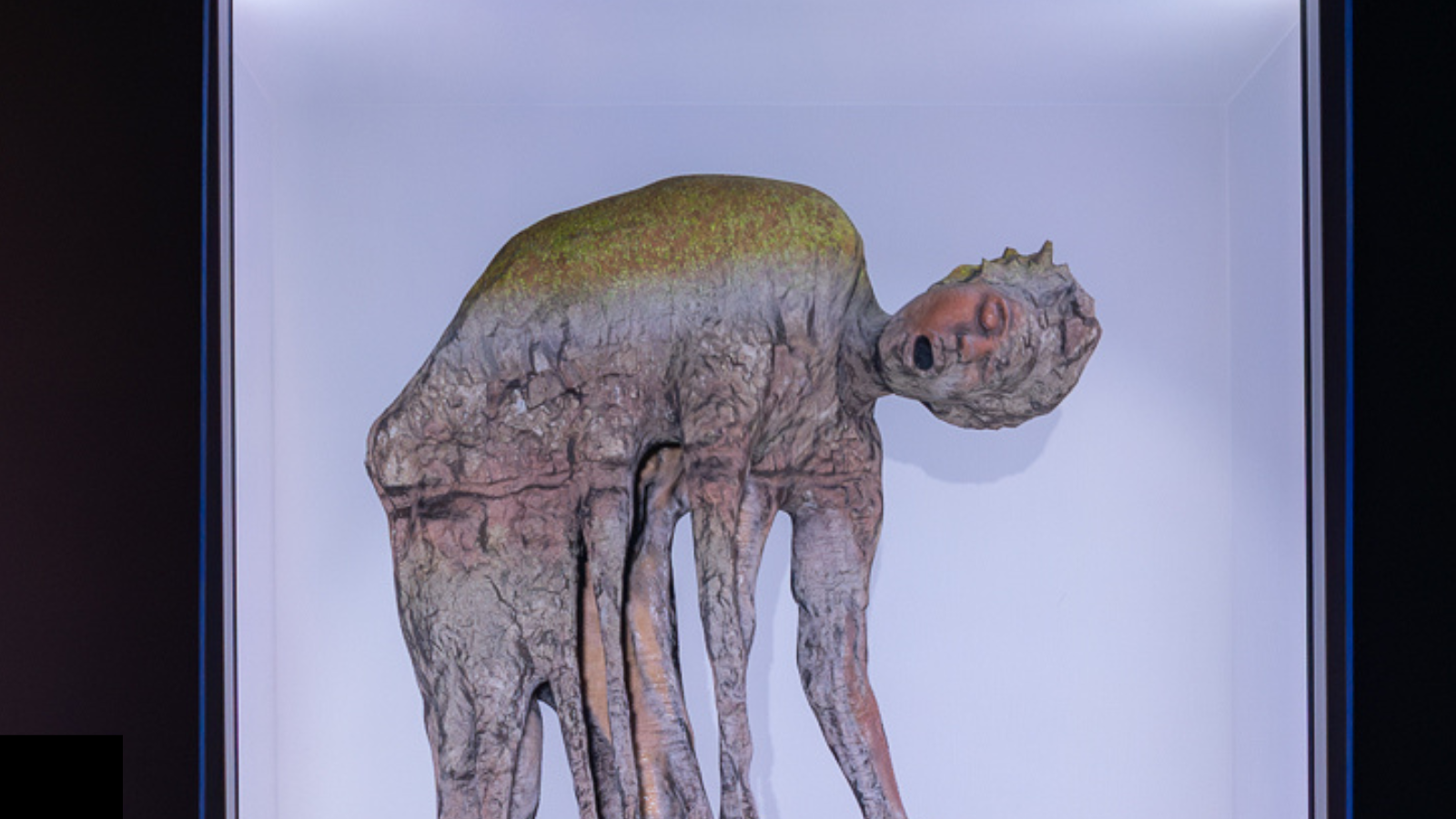 Detail of a 3-D digital scultpure presented on a transparent screen. A figure that is bending over with arms lowered towards their feet tilts their head toward the view.