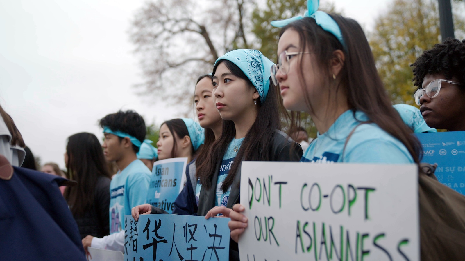 A group of students wearing light blue head gear and t-shirts hold up signs at what appears to be a protest