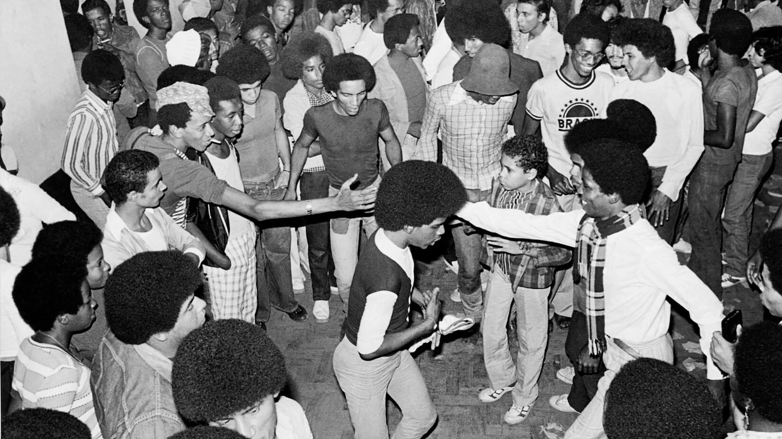 A black and white image of a group of people wearing afros dancing together joyously in a circle.