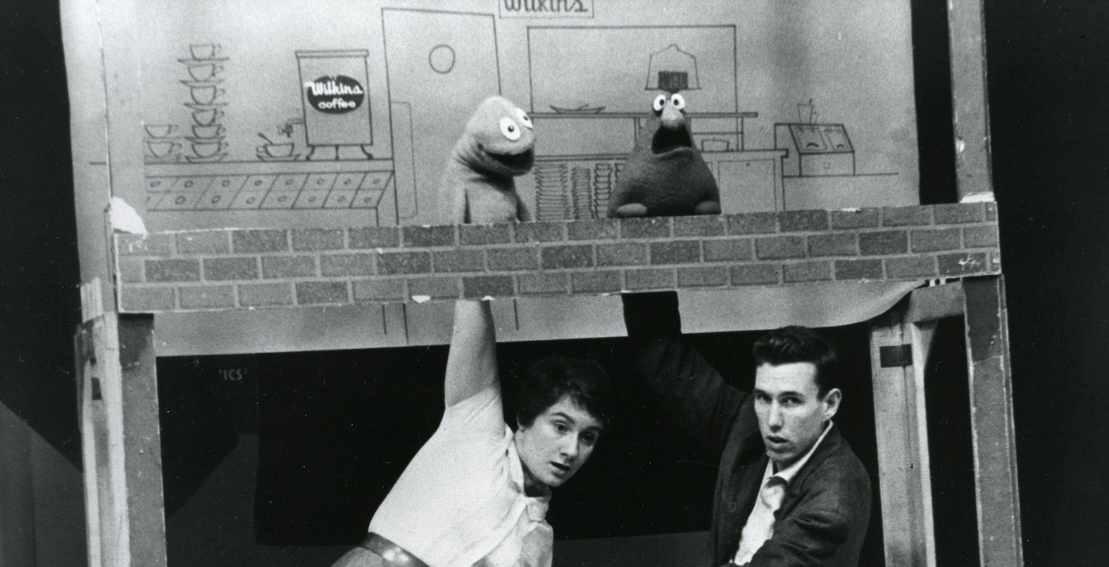 Two people crouch under a stage with their hands extended upwards holding silly looking puppets in a black and white photo from the 1950s.