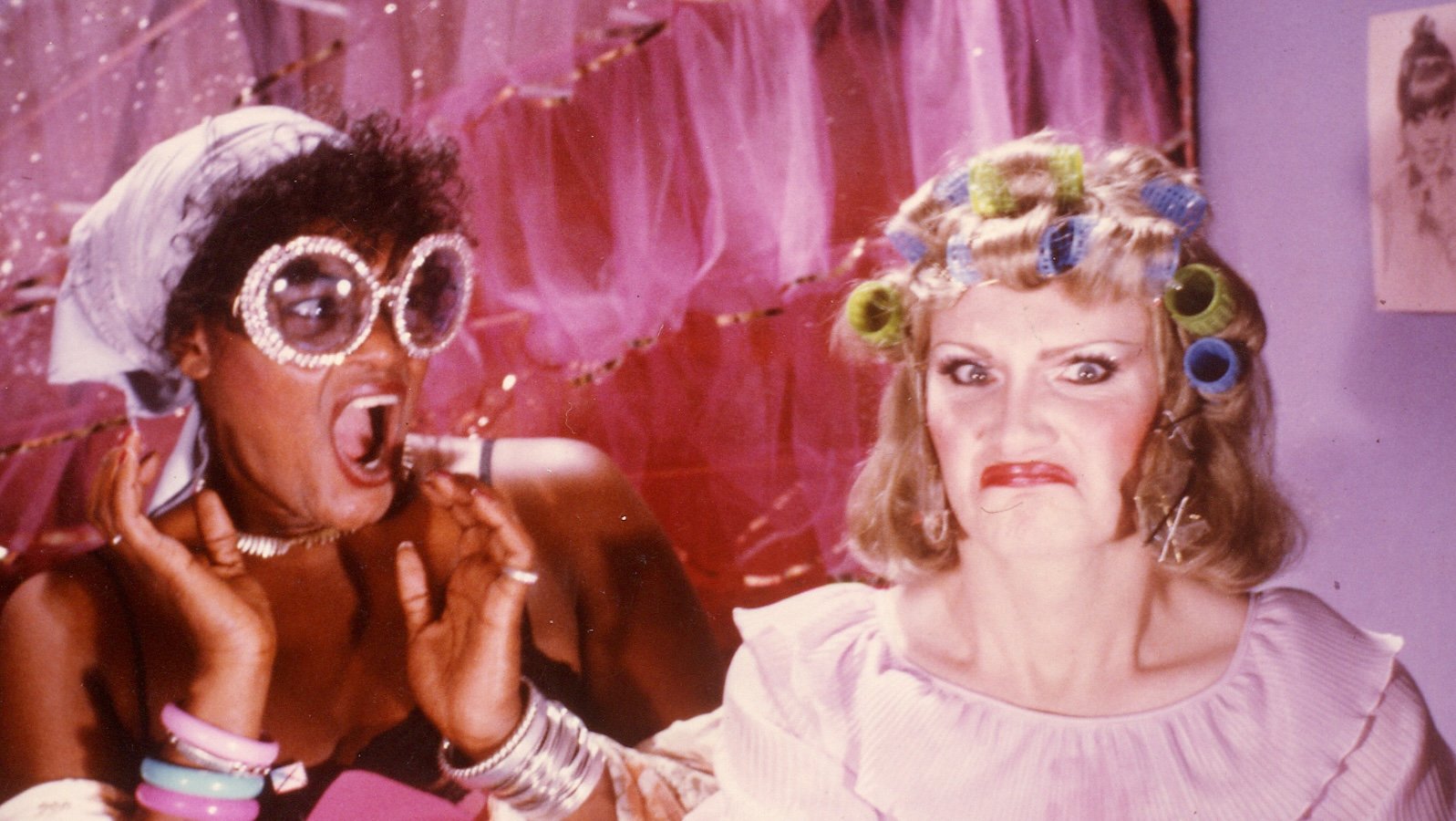 Two people against a pink, campy background: a dark haired woman with sunglasses making an outrageous shocked face with her mouth open on the right, and a blonde woman in hair rollers making a goofy face with pursed lips on the right