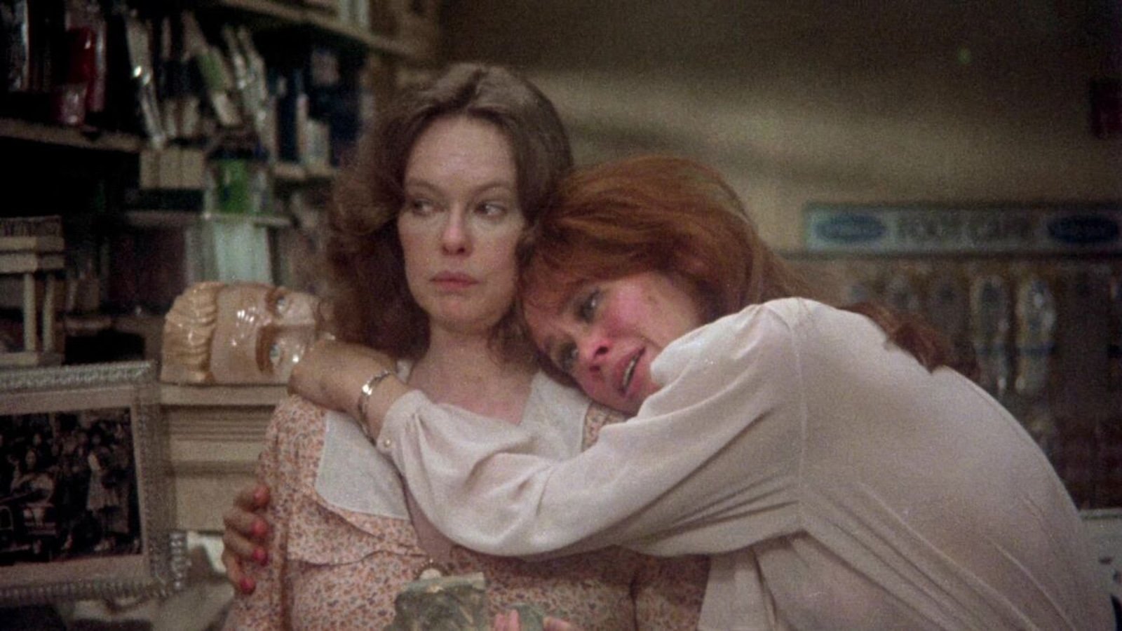 A redheaded woman on white puts her arms around the shoulders of a dark-haired woman in white