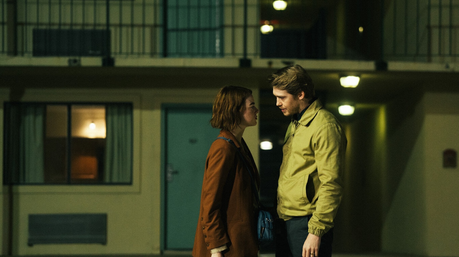 A woman in a burgundy coat and short reddish hair and a blond man in a crumply beige members-only jacket face each other intensely on the street in front of a motel.